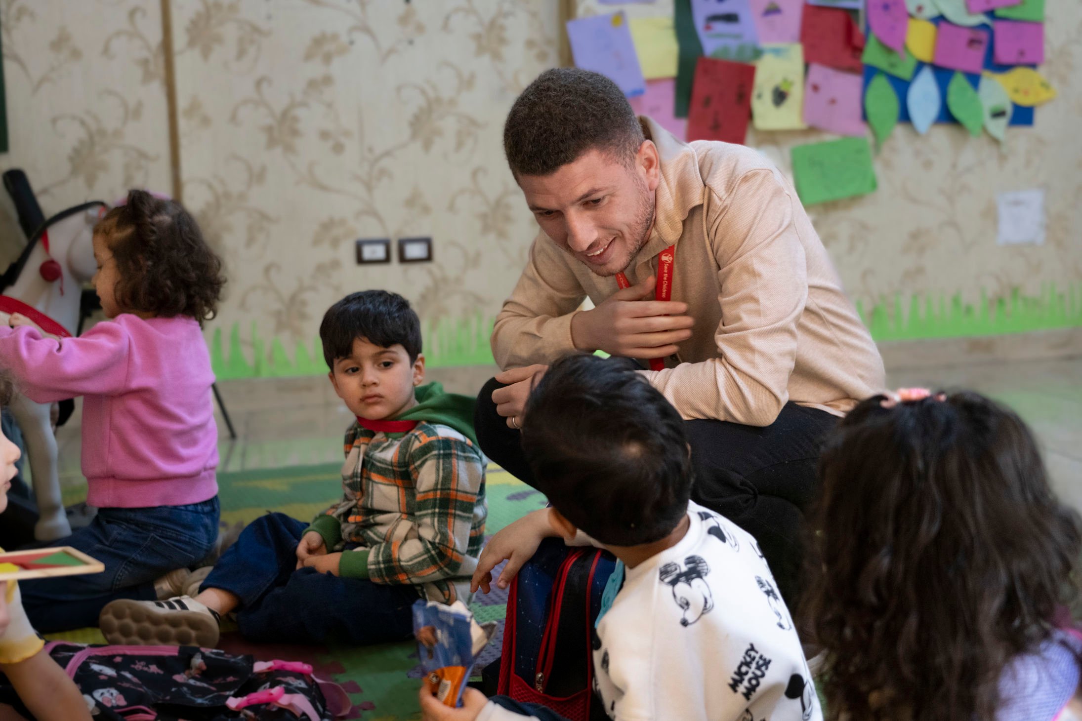 The image shows a group of children sitting on the floor in a colorful and playful environment, likely a classroom or activity space. An adult is crouched down, interacting with the children, wearing a light beige shirt and a red lanyard
