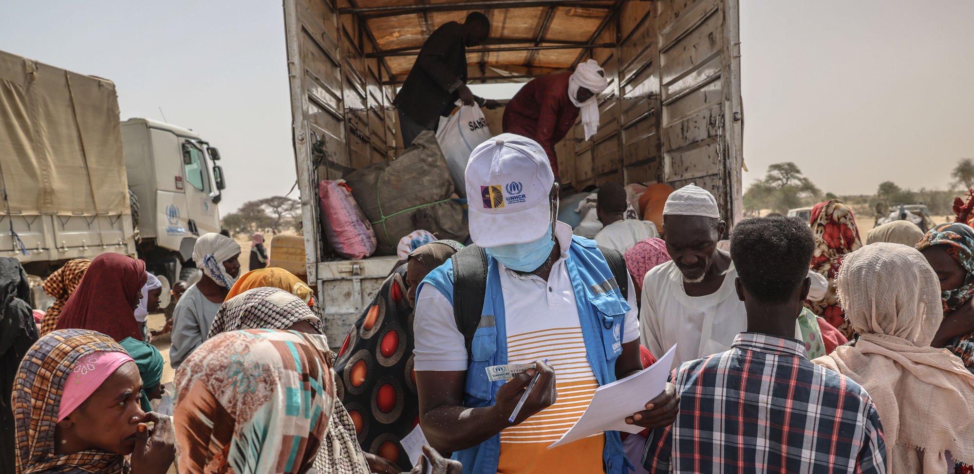 The image shows a humanitarian aid worker from UNHCR recording information from a group of people gathered around a truck. The truck is loaded with supplies, and the people are dressed in colorful clothing and head coverings. The scene takes place outdoors in a dry area