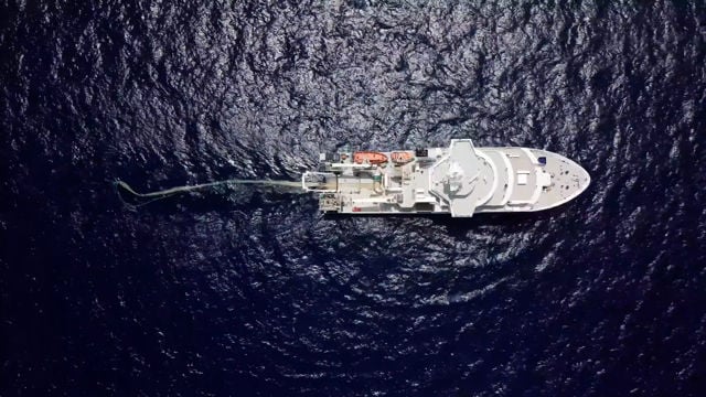 Bildet viser et skip sett ovenfra, midt ute på åpent hav. Skipet er stort og hvitt, med flere detaljer som skiller det ut, som ulike strukturer på dekk og livbåter. Havet rundt skipet er mørkeblått og glitrer i sollyset, med små bølger som gir en teksturert overflate.