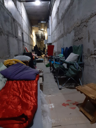 Dette bildet viser en lang, smal gang med betongvegger og et uferdig utseende, som minner om en kjeller eller en tilfluktsbunker. Langs begge sider av gangen står feltsenger, madrasser, soveposer og sammenleggbare stoler. Noen steder ligger det tepper og puter, og det er også små bord og personlige eiendeler plassert rundt. Belysningen er sparsommelig, med et par lyspærer i taket. Litt lenger inn i gangen sitter flere personer samlet, men de er litt uklare og i bakgrunnen.