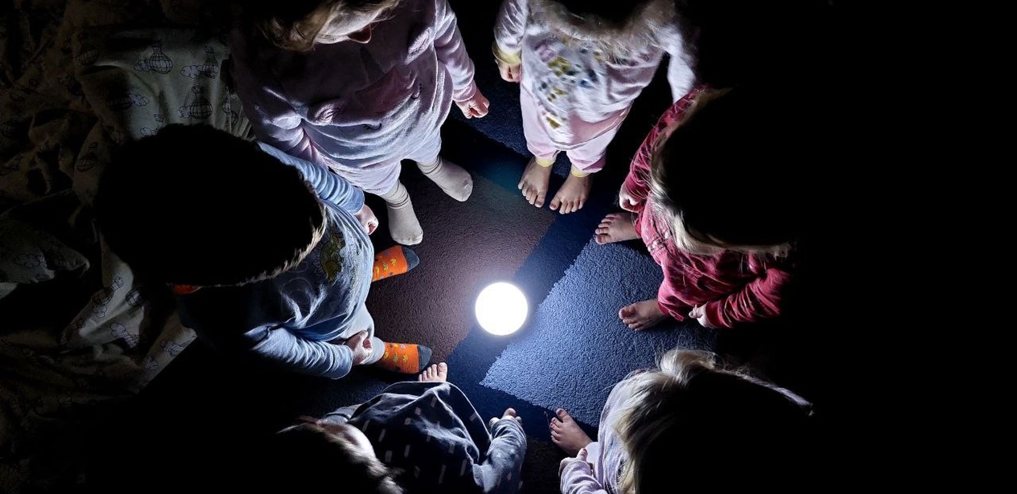 In the image, six small children are standing in a tight circle around a glowing orb or lamp on the floor. The photo is taken from above, showing the children's heads and shoulders, with their feet pointing toward the light. The room around them is dark, and the only illumination comes from the orb in the center, lighting up the children's faces and toes.