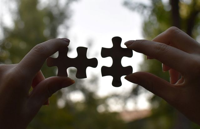 The image shows two hands, each holding a puzzle piece. The hands are facing each other, and the puzzle pieces are positioned so that they almost fit together, but are still slightly apart.