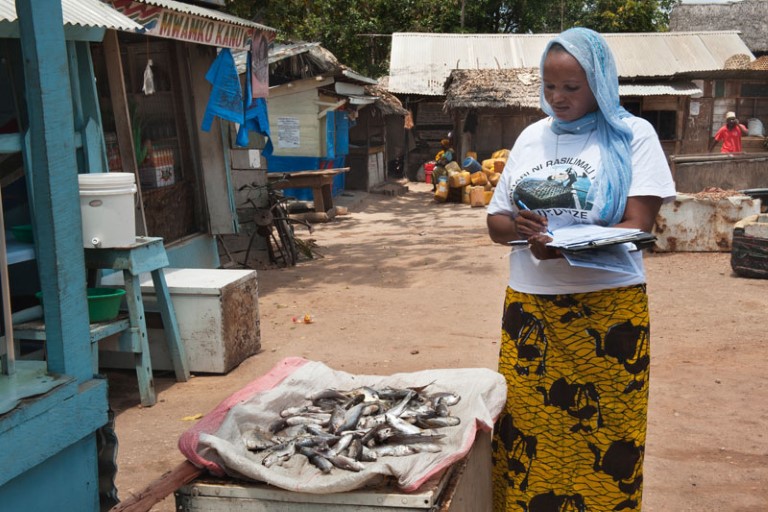 Kvinne står og skriver i en bok. Et fat med småfisk dominerer bildet. Tanzania