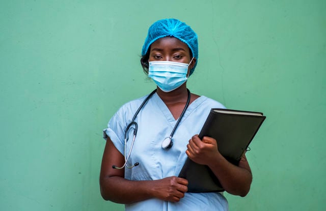 Nurse in blue scrubs with a stethoscope around the neck holding a black binder, standing against a green wall.