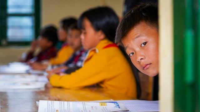 Several children sit at desks in a classroom, with the focus on a boy looking out the window.