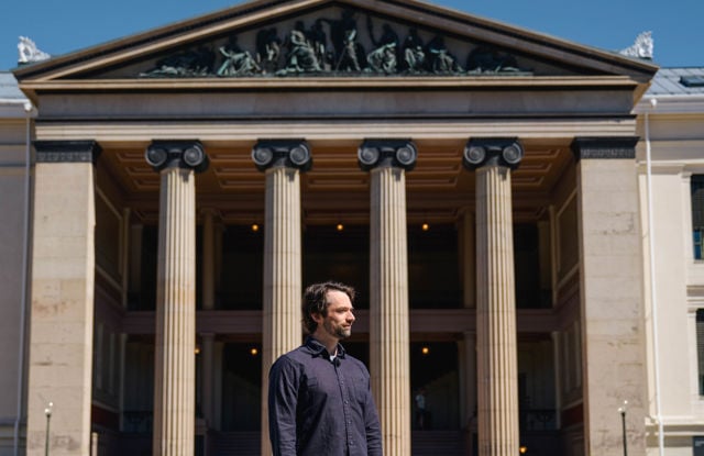 A man stands in front of a large building with columns and a triangular pediment. He looks to the side, while the building fills most of the image.